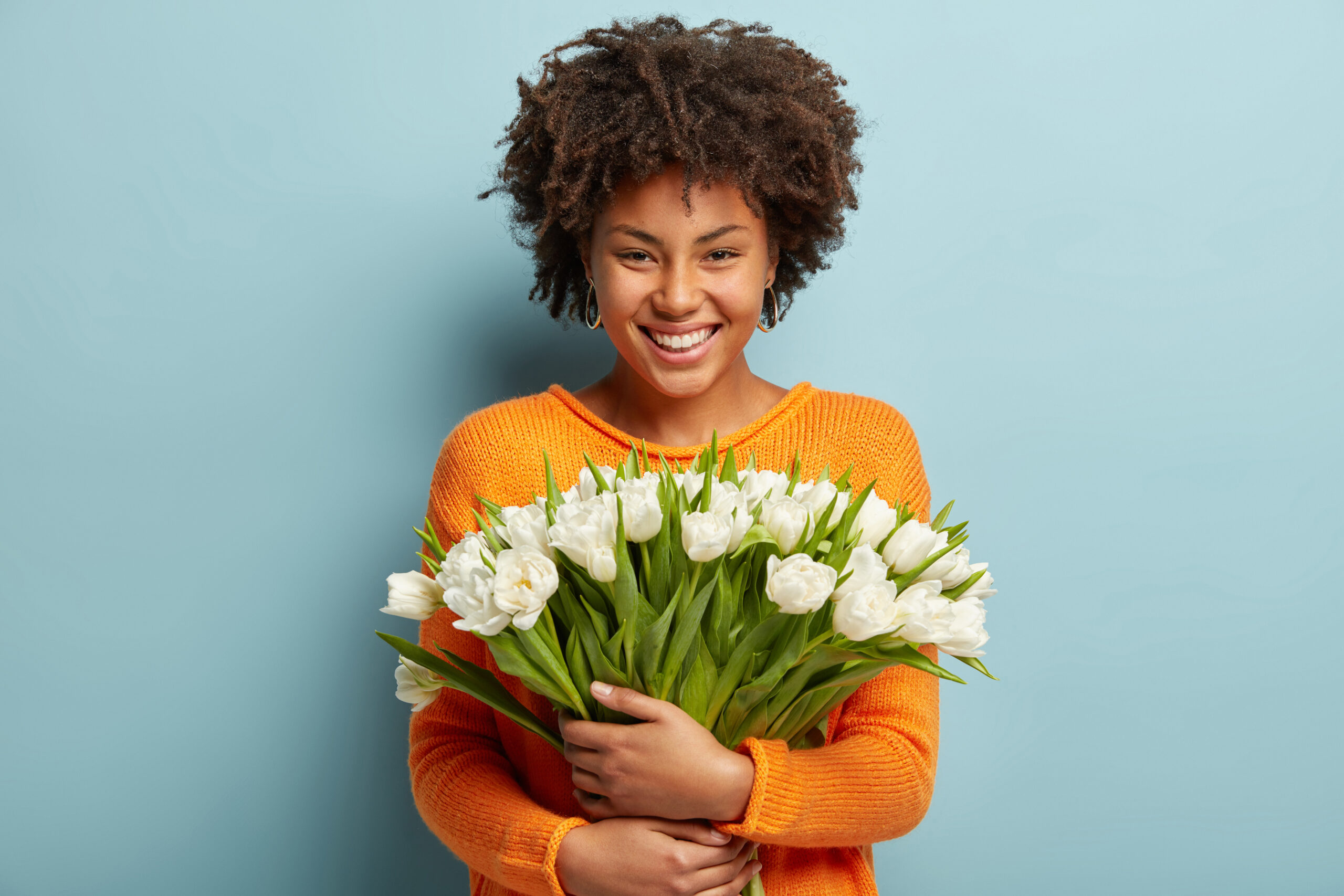 Indoor Shot Of Beautiful Smiling Young Woman With Afro Haircut Holds Big Bouquet Of Spring White Flowers, Rejoices Coming Womens Day Soon, Dressed In Orange Jumper, Isolated Over Blue Background.